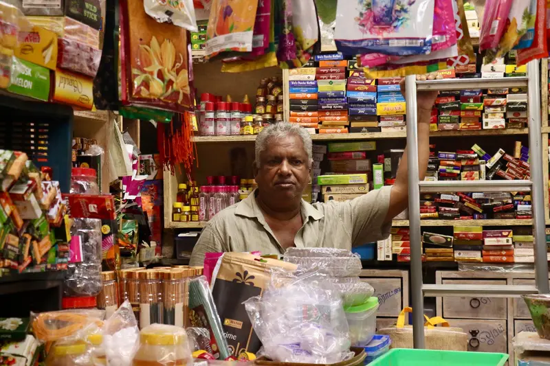 A shopkeeper behind a counter in a colourful general store packed with goods (illustrative photo)